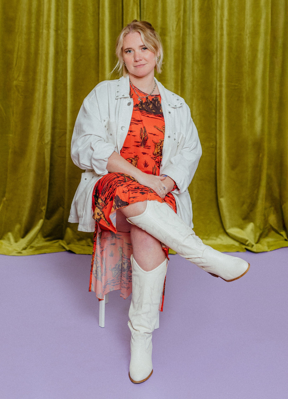 Woman in a red dress and white jacket sitting against a green curtain on a purple floor.