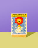 A double print pack of novelty stickers by The Mask Press, featuring a cute lion face and the text 'PLEASE DON'T PICK ME' on a light blue textured background. The pack is displayed on a wooden stand against a split purple and yellow background.