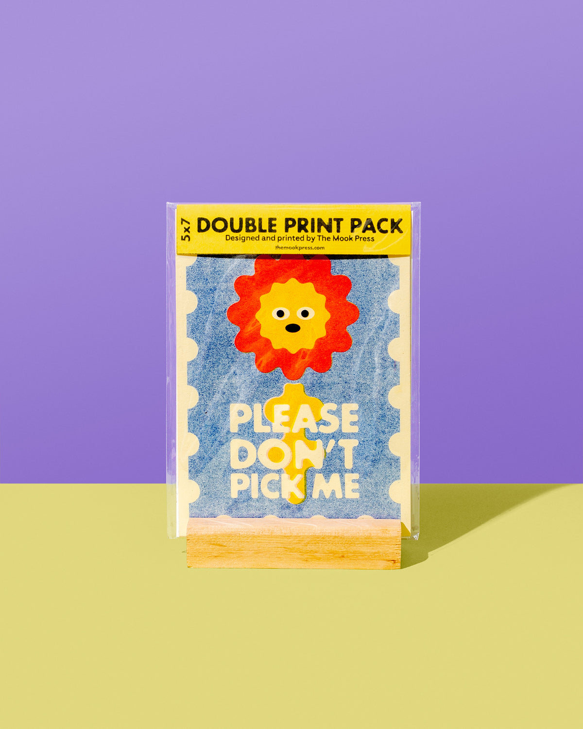 A double print pack of novelty stickers by The Mask Press, featuring a cute lion face and the text 'PLEASE DON'T PICK ME' on a light blue textured background. The pack is displayed on a wooden stand against a split purple and yellow background.