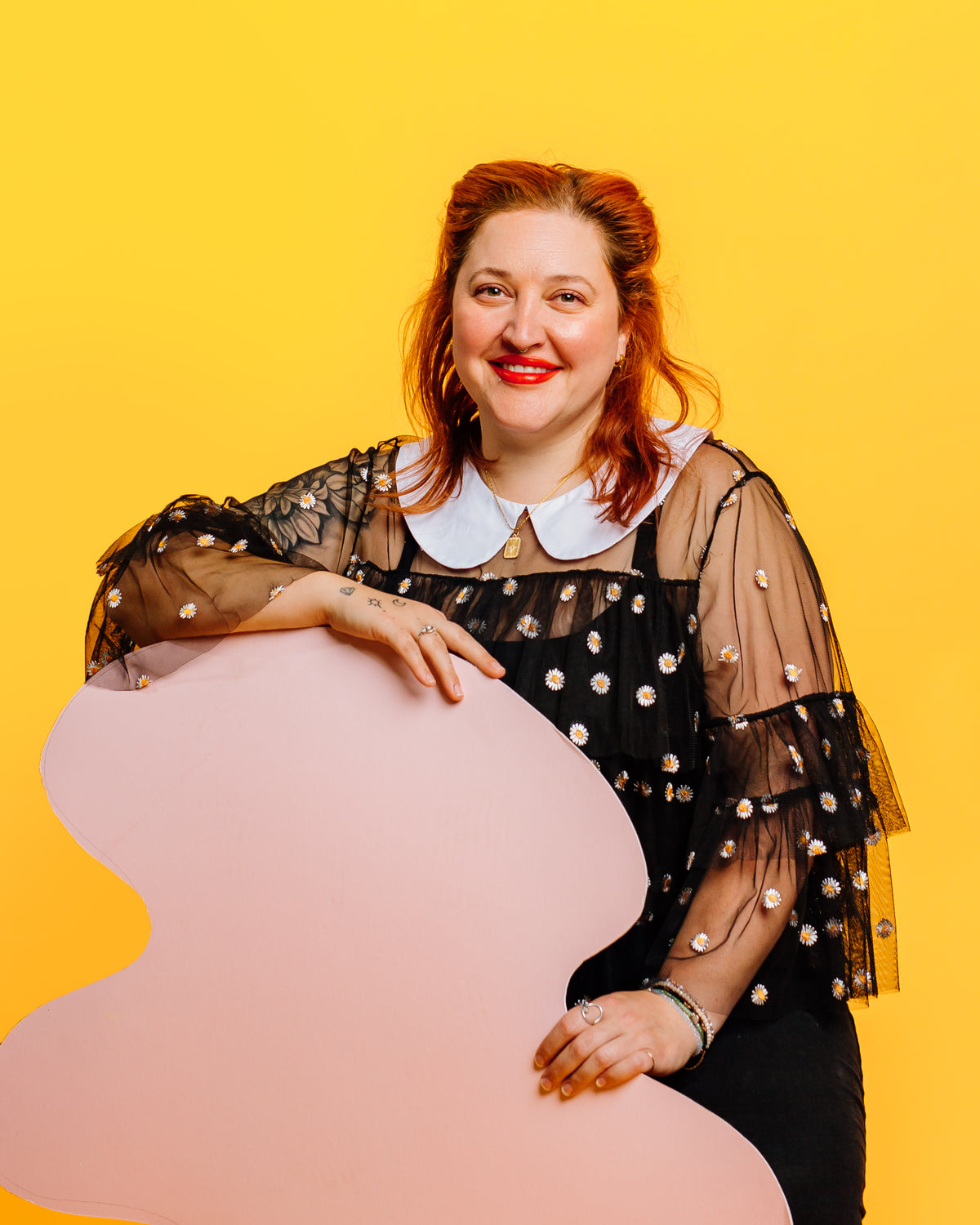 Woman with red hair, red lipstick holding pink shaper in front of yellow background. Arkansas Local Concrete Artist sells work at Rogers Arkansas Best gift shop Kaleidoscope Collective
