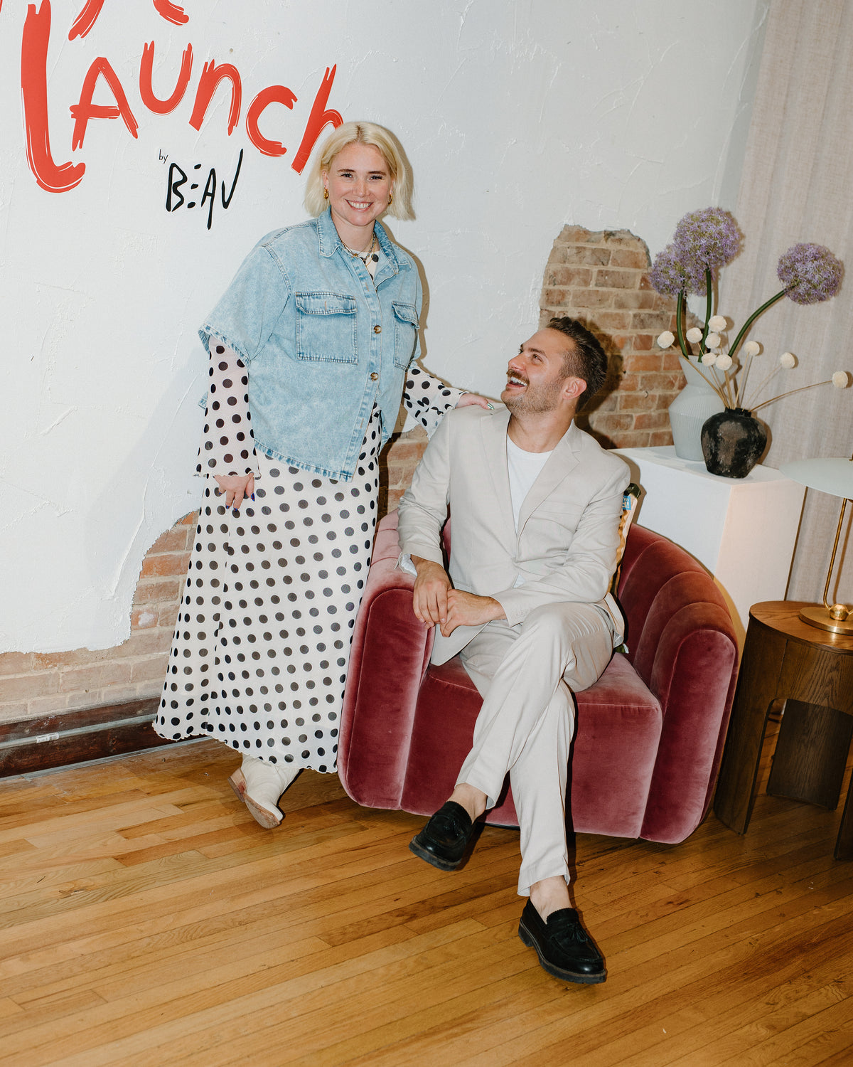 Woman in dotted dress with man sitting in char with text on a wall and a hardwood floor in art gallery laughing