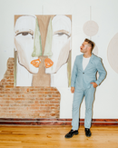 Man in blue suit looking at a painting he made with two faces hanging on plaster and exposed brick wall in downtown rogers, arkansas