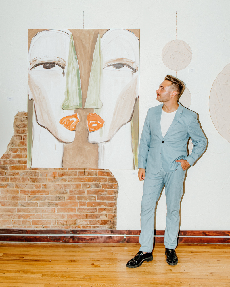 Man in blue suit looking at a painting he made with two faces hanging on plaster and exposed brick wall in downtown rogers, arkansas