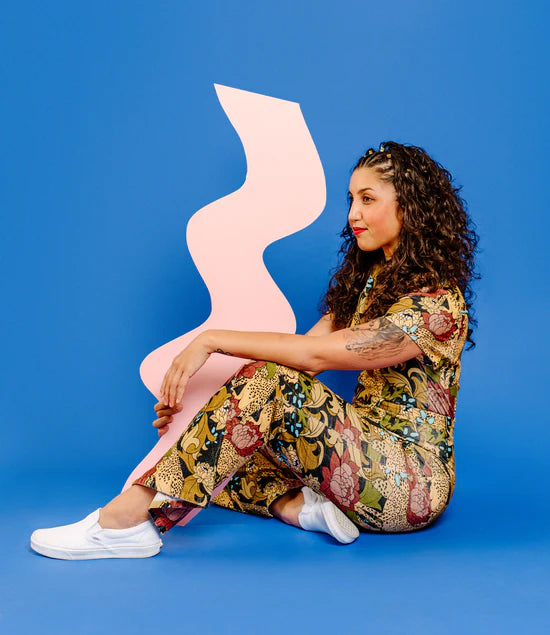 Creative Artist with curly hair in front of bright shape background with creative jumpsuit - embroidery artist and creative teacher in Northwest Arkansas