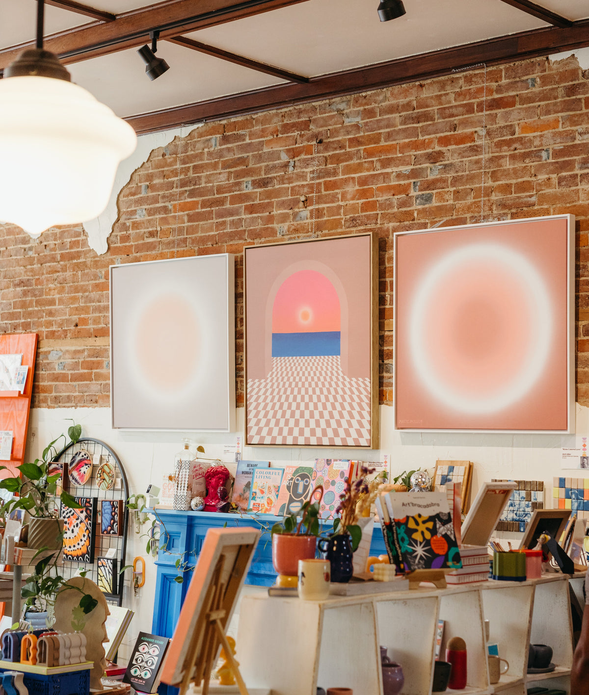 A styled shop filled with local art with exposed brick walls, three large abstract art prints hanging above a blue fireplace, shelves with books and decor, and a person partially visible on the right side.