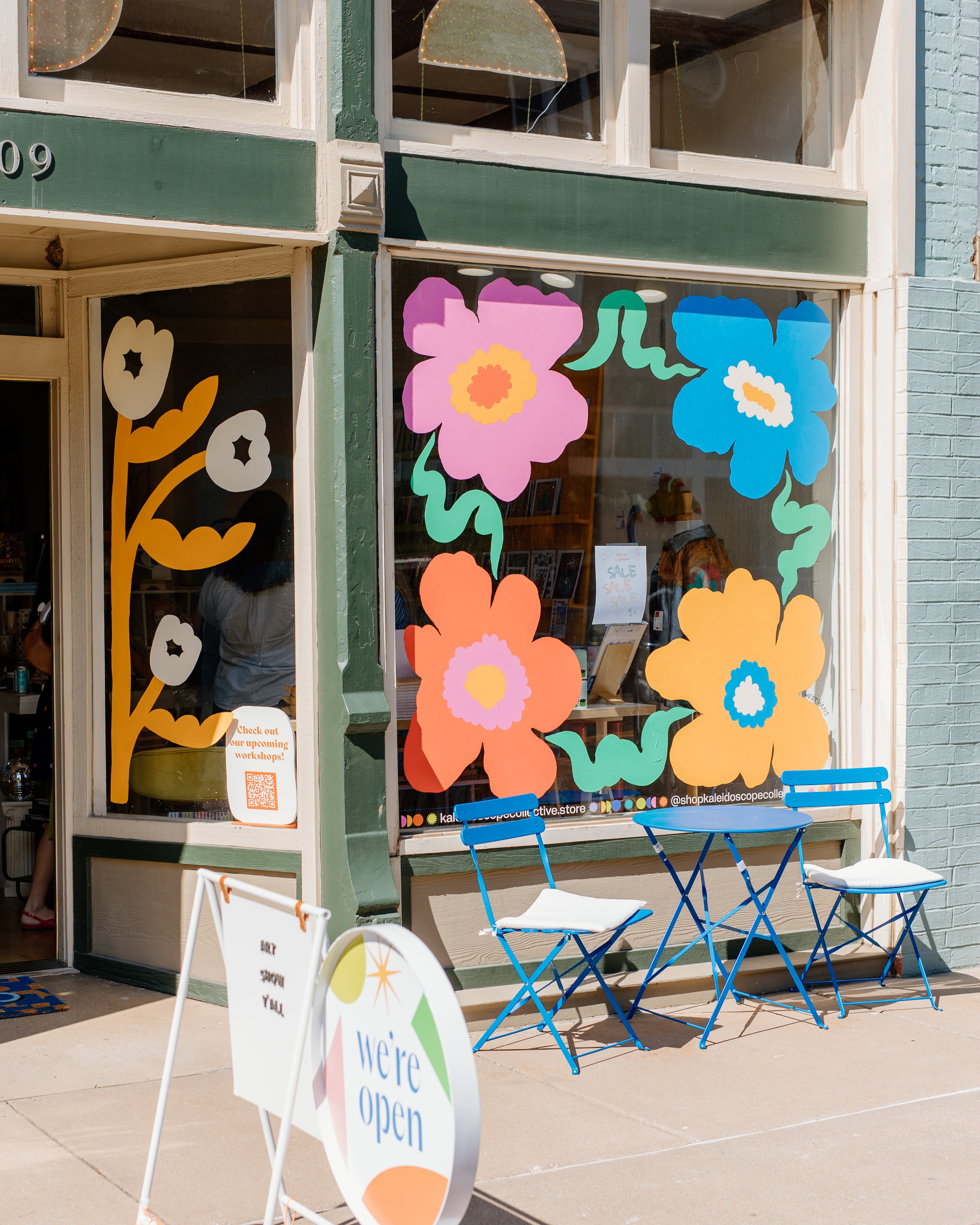 A storefront window decorated with large colorful paper flowers in pink, orange, blue, yellow, and green, with two blue folding chairs placed outside on the sidewalk. A round sign on the sidewalk reads 'we're open'.