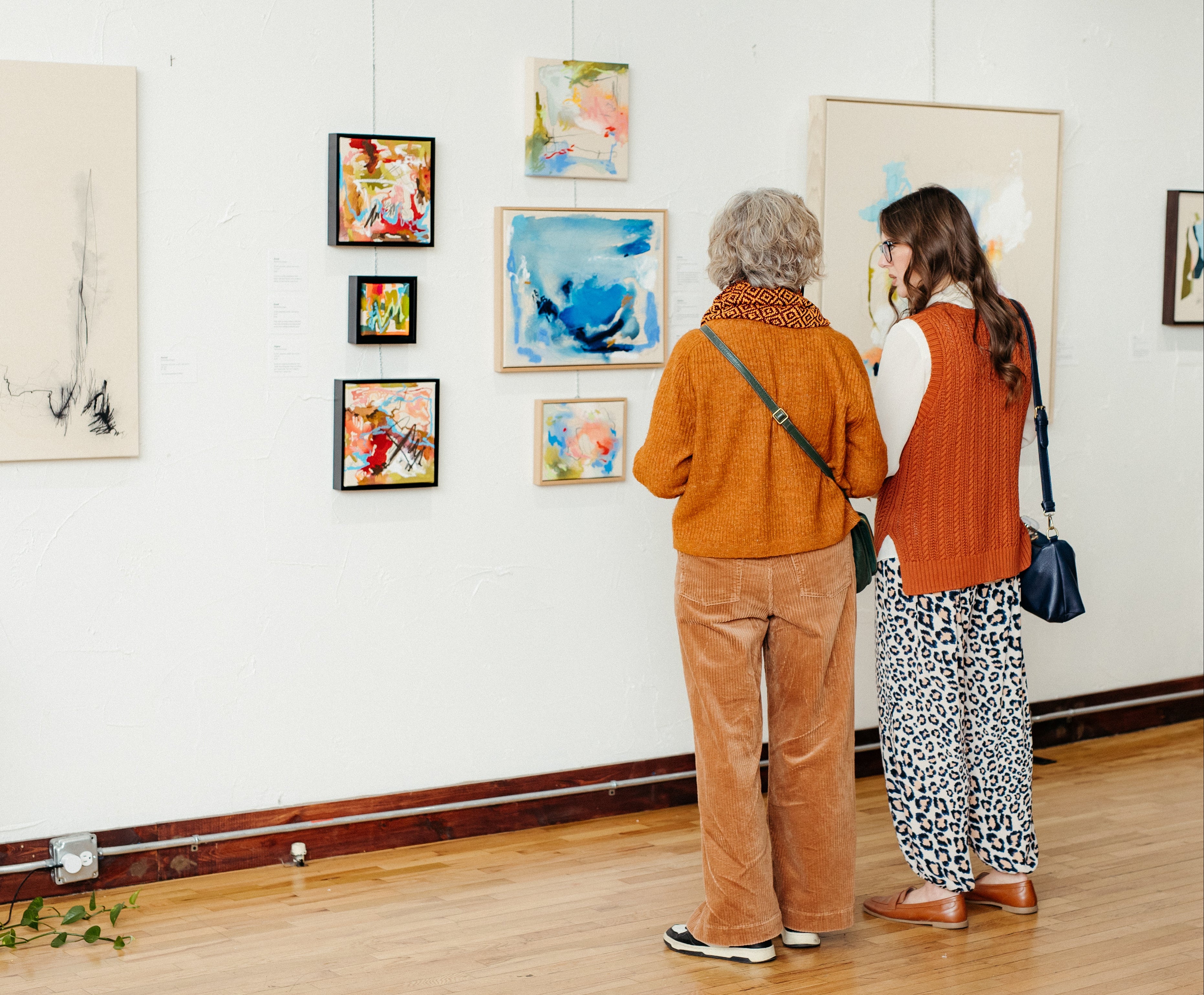 Two women viewing framed abstract paintings on a white gallery wall in a bright art gallery with wooden floors at Kaleidoscope collective in Rogers, Arkansas