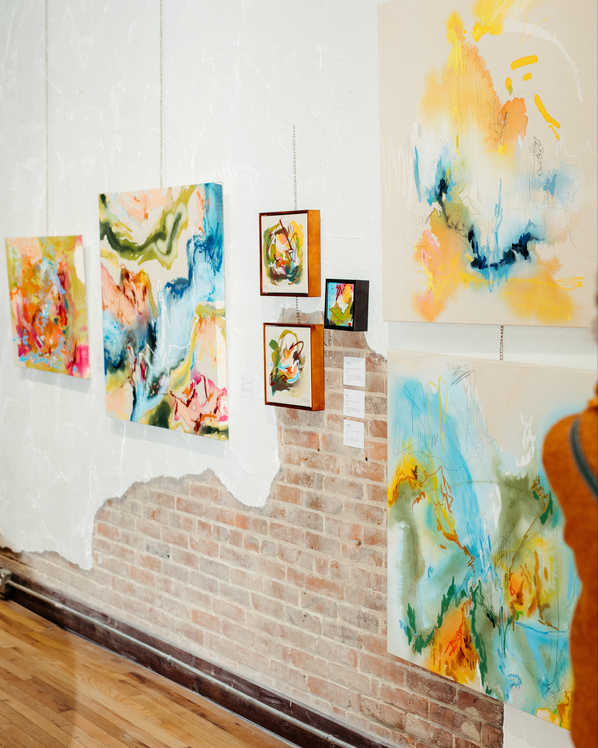 A gallery-style interior showing several colorful abstract paintings hanging on a white wall above a brick baseboard and wood floor. The artwork features blue, yellow, orange, green, and pink tones, with one large abstract piece on the right and smaller framed pieces in the center.