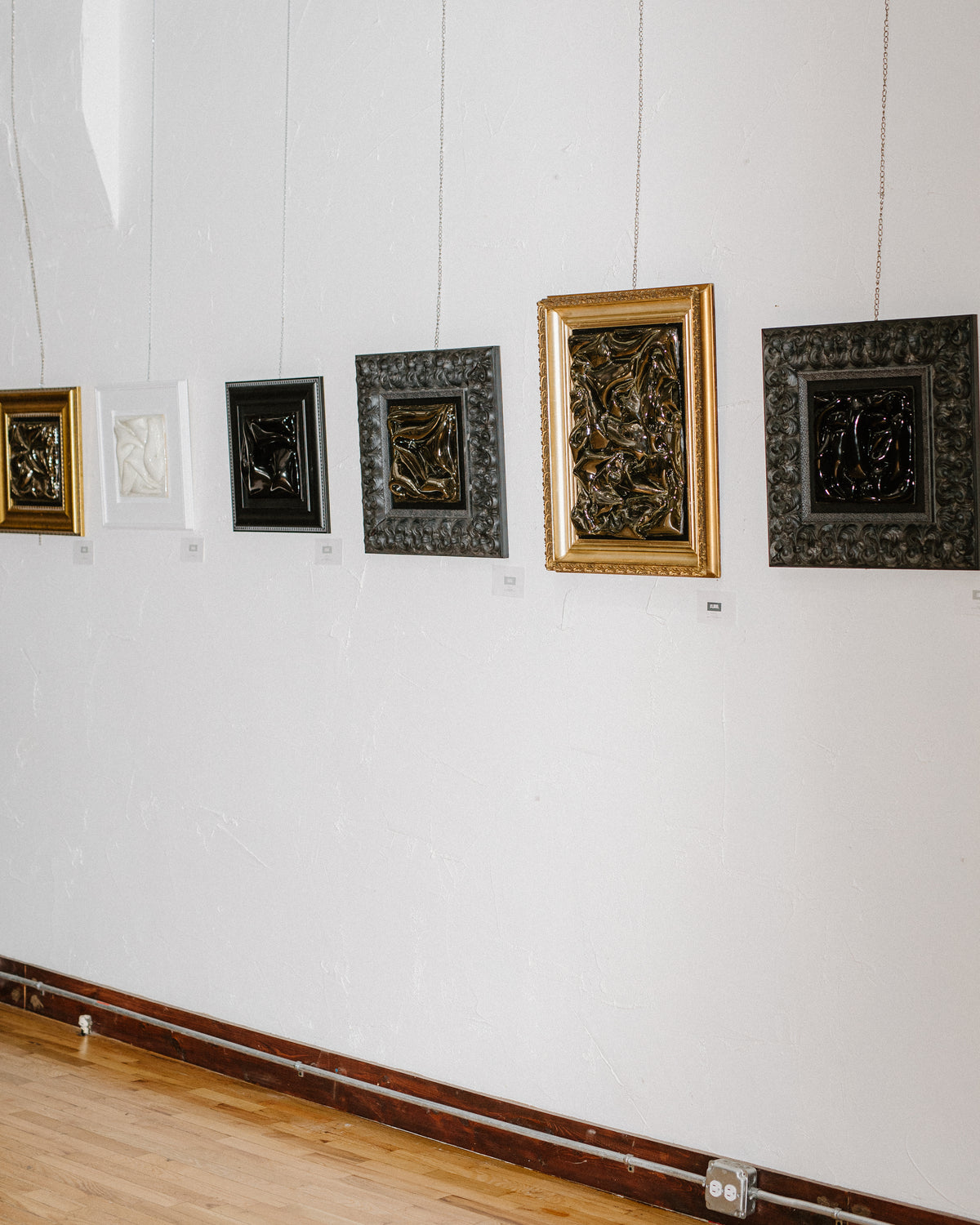 Six framed decorative wall art pieces displayed in a gallery or exhibition setting, hung at varying heights on a white wall above a wooden floor.